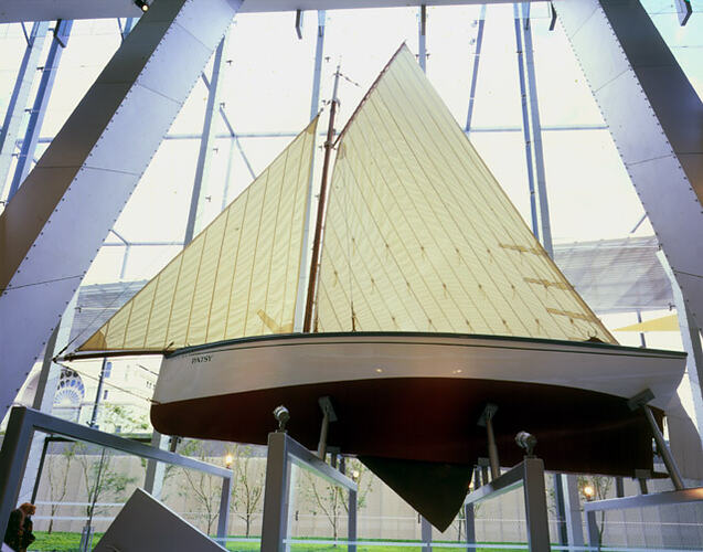 Boat with white sail suspended inside buildign from roof. Glass wall behind.