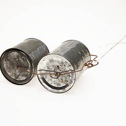 Two metal tins with lids. Bound together with wire. Cord at one end to pull them along. End view.