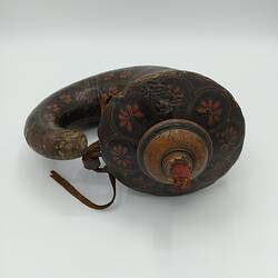 Top view of volute shaped wood flask covered with red patterned papier mache. Wide sealed opening with wooden stopper curls to thin spout. Has strap.