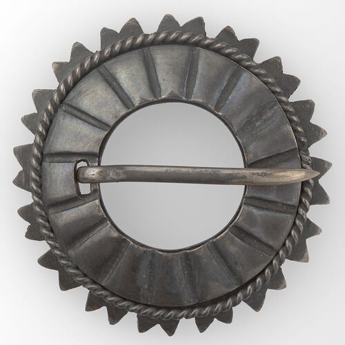 Ring-shaped metal brooch with radiating grooves and zigzag edge. Pin across middle.