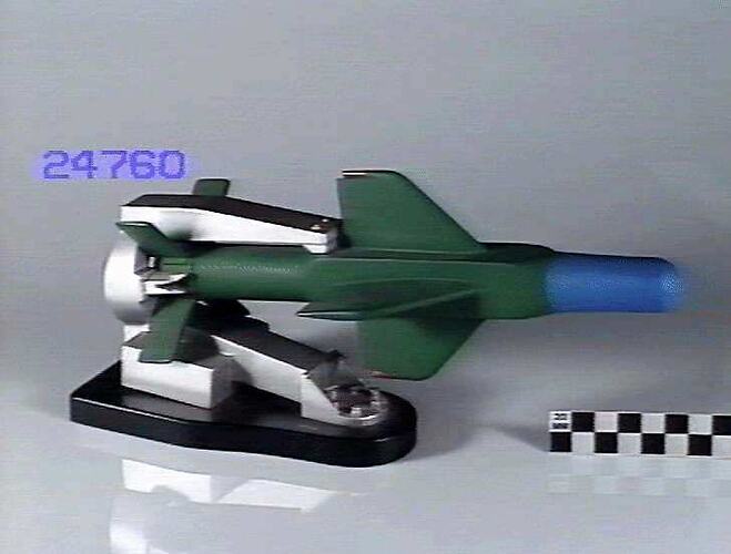 Model missile. Four vertical and four cruciform wings, nose is painted blue, body and wings are green. Support is silver, base is black.