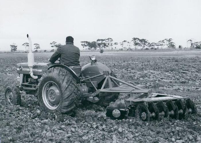 Rear left side view of a tractor coupled to a disc harrow in a ploughed field.