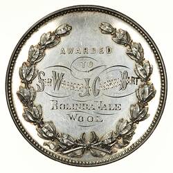 Round medal with decorative text framed by ornate laurel wreath.