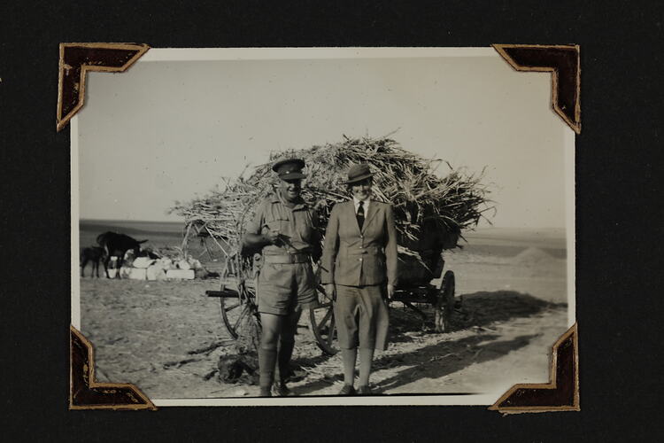 Woman and man in uniform standing in front of wagon large holding bale of hay, two donkeys in left background.