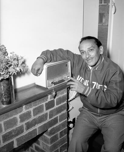 Man sitting beside a brick fireplace with his arm around a radio on the mantelpiece.