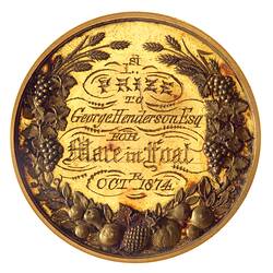 Round gold medal with decorative engraved text framed by floral and fruit wreath.