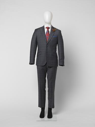 Grey suit jacket and pants with blue line large check pattern. White shirt, red patterned tie. Brown feather corsage, blue patterned kerchief strip at pocket.