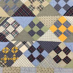 Detail of colourful blue/grey/yellow/green patchwork quilt. Brown trim.