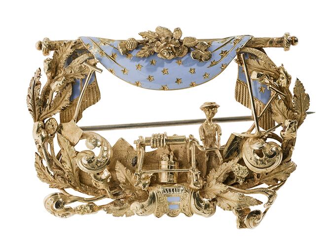 Rectangular gold brooch. Ornate detail, open centre. Blue enamel drapery at top. Miner, tools and foliage.