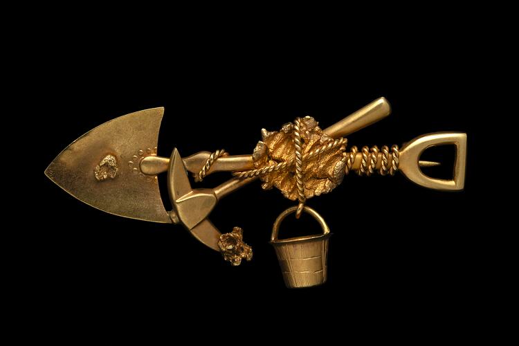 Gold brooch with pointed shovel, pick and bucket bound with rope around a larger nugget in centre. Small nugget on shovel and pick end.