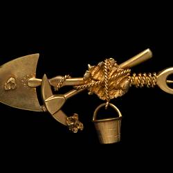Gold brooch with pointed shovel, pick and bucket bound with rope around a larger nugget in centre. Small nugget on shovel and pick end.