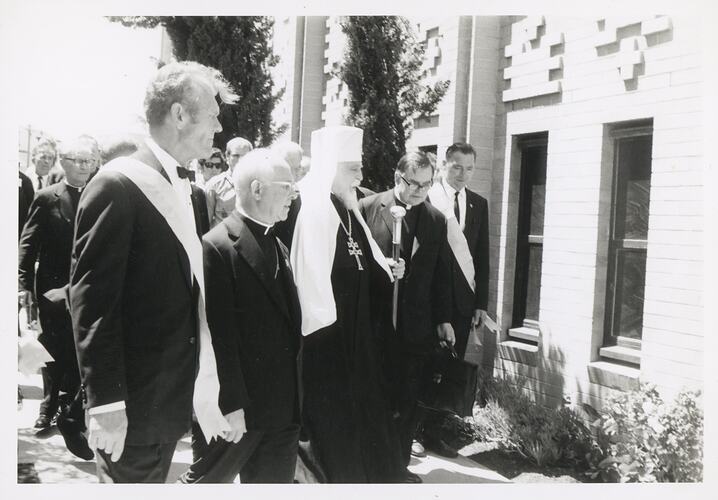 Cardinal, bishop and other men outside a brick building, possible walking along side it or about to enter.