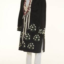 Black coat with white floral detail over white chinos pants. Long beaded necklaces.