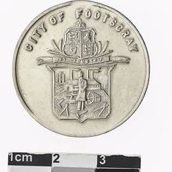 Round silver coloured medal with coat of arms and text above.