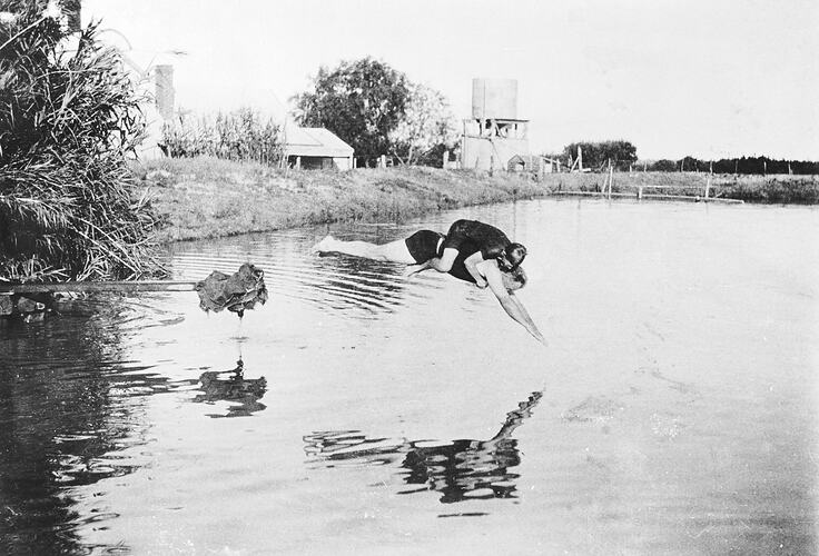 A man diving into a dam with a boy on his back. There are farm buildings and a water tank in the background.