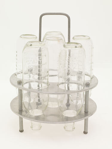 Circular metal rack with handle holding 6 upturned glass babies' bottles.