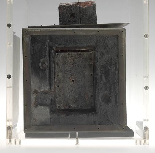 Black metal cube-shaped box (burnt) in clear perspex box. Rectangular panel at front and section on top.