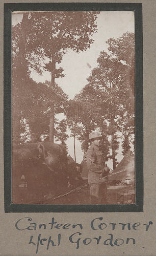 Soldier in uniform standing in camp ground with horse to the left and tents and trees in background.