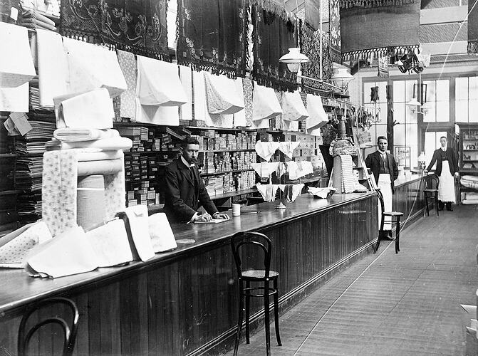 Three men stand behind and along a long wooden service counter at left. Fabric fills shop.