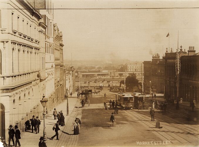 City street with stone office buildings, a railway bridge, and several factory complexes in the distance. Two cable trams and horse drawn vehicles in street.