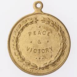 Round gold coloured medal with text surrounded by wreath.