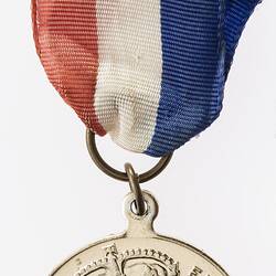 Round medal with profile of crowned woman and text surrounding, with red, white and blue ribbon.