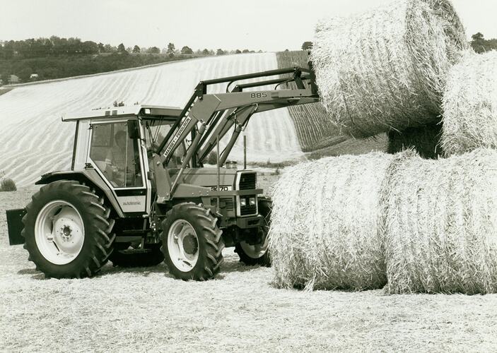 Tractor fitted with a front end loader round hay bale attachment stacking round bales in a field.