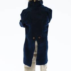 Miniature man in dark coat, light pants. Has a hat and fair hair. View from back.