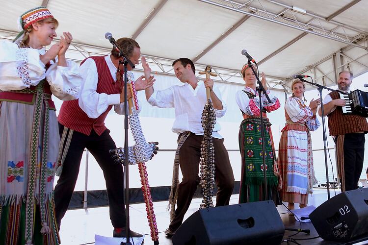 Six people in Lithuanian costume perform on stage. They sing, clap, play an accordion and two largerphones.