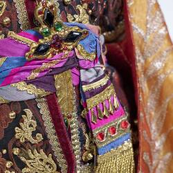 Detail of bejewelled ornate burgundy tunic with blue and purple belt and orange coat with gold embroidery.
