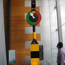 Man looking up at yellow and black striped pole with green and red round dial near top.