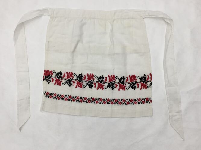 White hessian cotton with long ties, red and black leaf and vine embroidered pattern across front lower section.