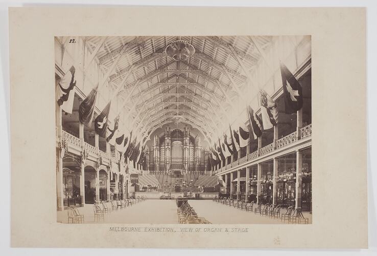 Interior of the Great Hall of the Melbourne Exhibition Building at the 1880 Exhibition, showing flags, chairs, displays of light fittings and other goods, the choir stalls and the organ. The view is looking west along the Great Hall from the Dome.