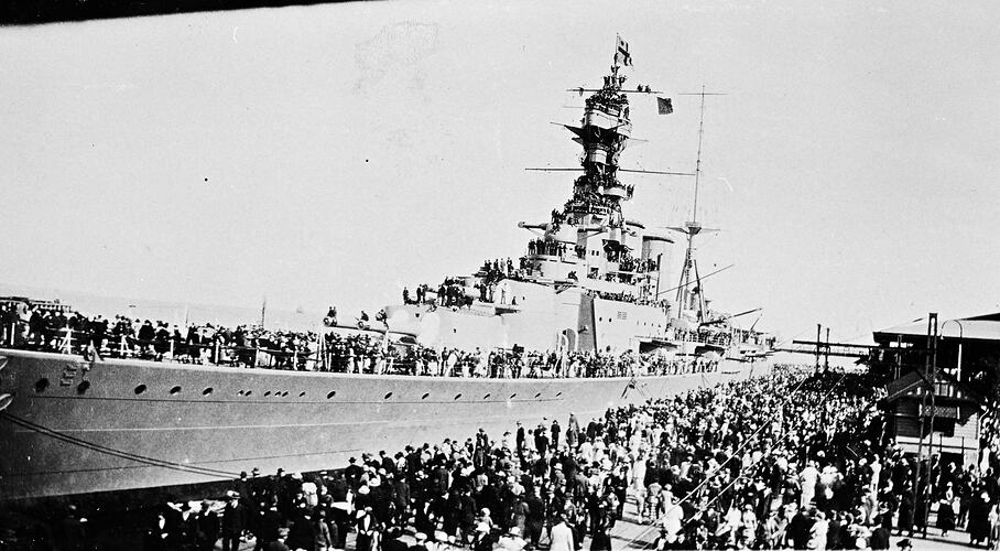 Crowd inspecting berthed warship from a pier and on board. Some are on high rigging and masts.