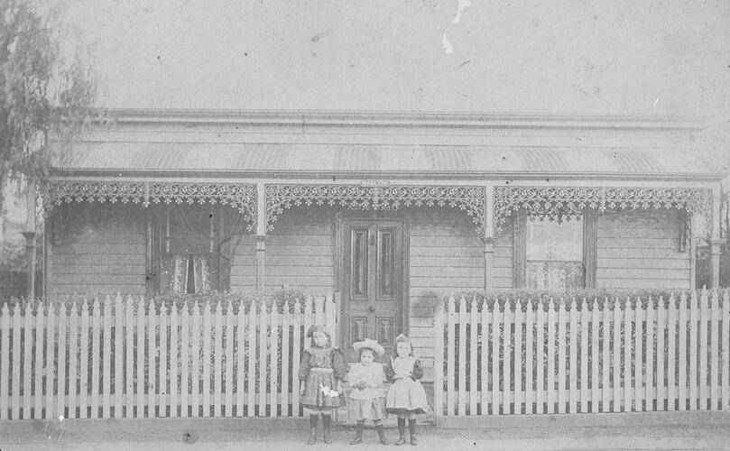Three small children in front of a weatherboard house with ironwork lacework verandah. Front picket fence.