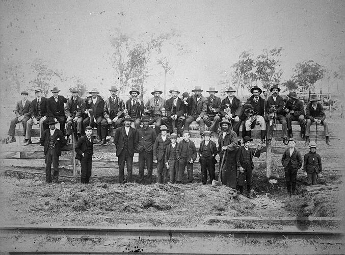 Group of stockmen and dogs pose on and in front of a fence.