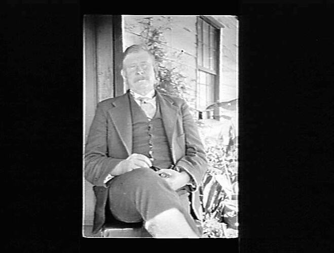 Photographic negative of man seated outside house.