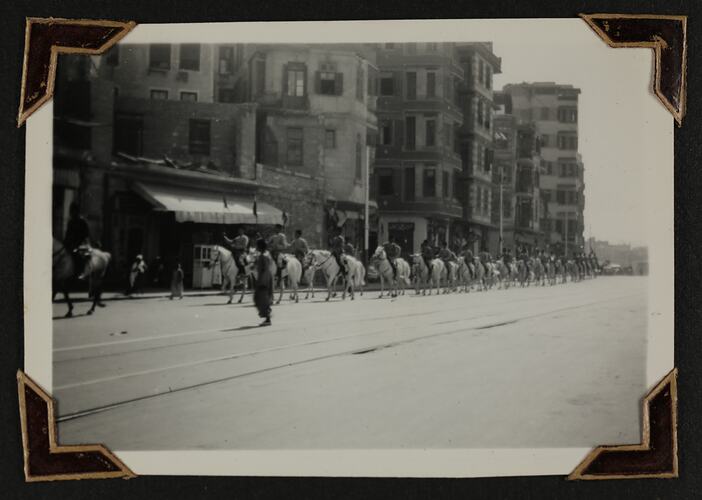 Line men on horseback walking down a street, tall buildings behind them.