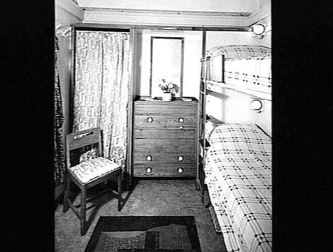 Ship interior. Bunk beds on right, chair on left. Chest of drawers in centre.