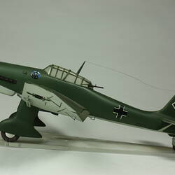 Model aeroplane. Dark green with black cross on tail. Black propeller and wheels. Left profile.