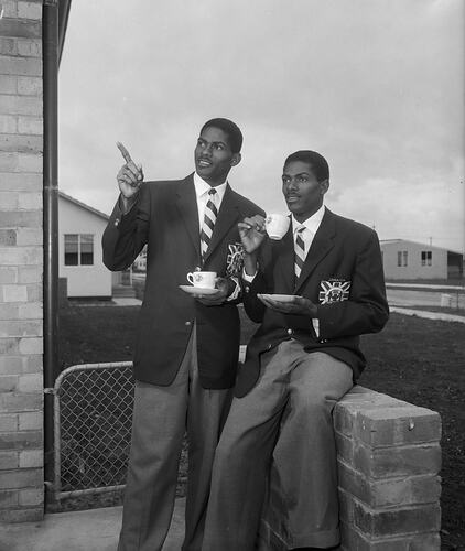 Two sportsmen wearing team blazers pose with cups of tea.