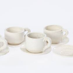 Model of six miniature white cups and saucers. One cup beside its saucer.
