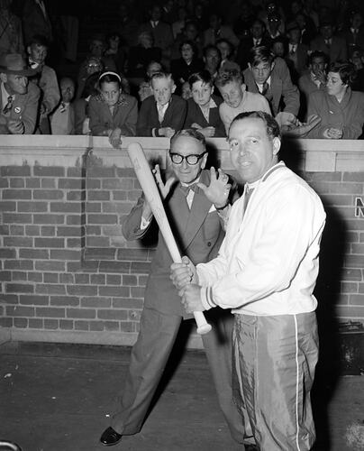 Man holding a baseball bat. Man behind has hands up as 'catcher'. Crowd behind them looks on.