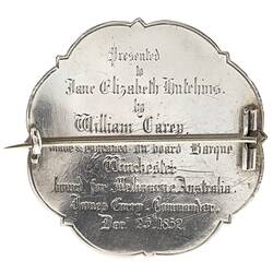 Back of brooch made from silver coin. Scrolled edge. Raised lettering. Pin across centre.