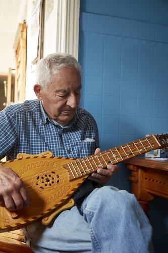 Man sitting at table playing the mandolin. Blue wall behind.