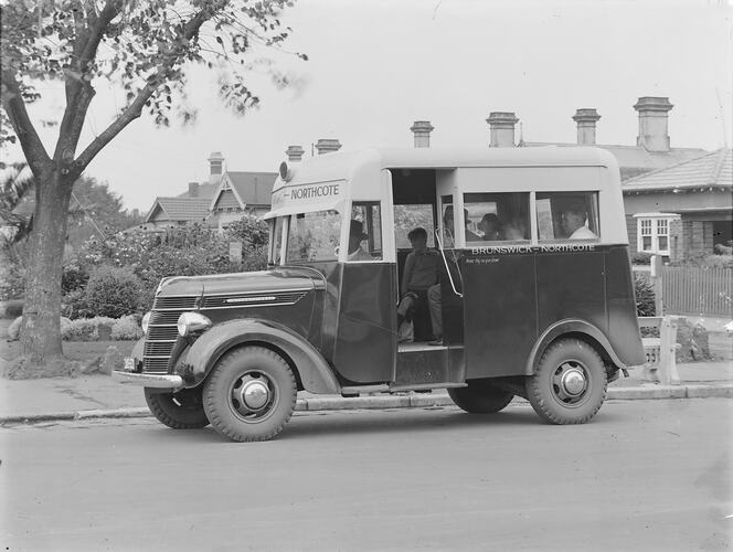 Small two-toned motor bus marked 'Brunswick-Northcote' stopped at roadside in front of residences and garden.
