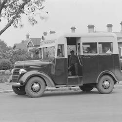 Small two-toned motor bus marked 'Brunswick-Northcote' stopped at roadside in front of residences and garden.