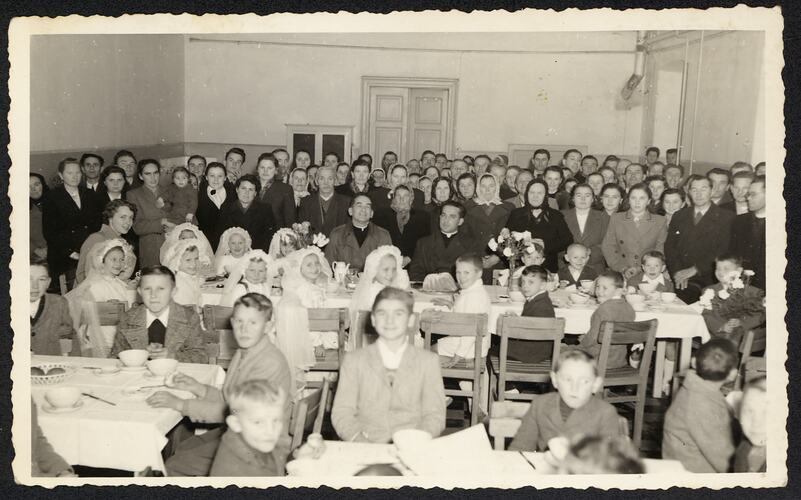 Large group of people sitting at multiple tables posing for photo. Some of the young girls wear white veils.