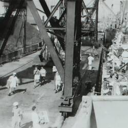 Sailors carrying wooden crates on the right, docked ship with sailors on deck on the right.