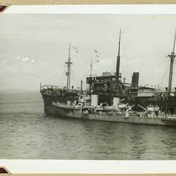 Small heavily damaged ship in water in front of larger ship.